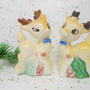 May include: A pair of vintage ceramic reindeer figurines. Each reindeer has a cream-colored body, brown antlers, and a blue bow tie. They have painted details including eyes, a red flower, and green leaves.