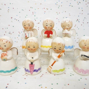 May include: A collection of eight ceramic angel figurines. Each angel has a white dress with a colorful trim, blonde hair, and small wings. The angels hold various items, including a heart, a book, and a broom.