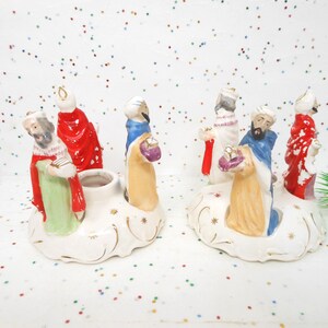 May include: Two ceramic nativity candle holders, each featuring three figures of the Three Wise Men. The figures are dressed in red, green, blue, and gold robes. The candle holders are white with gold accents.