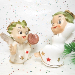 May include: Two vintage porcelain angel figurines with green hair and wings. One angel is holding a heart-shaped object and the other is sitting with its arms behind its head.