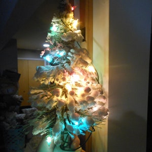 May include: A small, artificial Christmas tree with white, snow-like flocking and colorful lights. The tree is decorated with small, round ornaments and is illuminated by a string of lights in various colors. The tree is placed indoors.