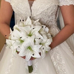 Puede incluir: Un ramo de novia de lirios blancos con centros verdes y estambres negros. El ramo se sostiene con una cinta blanca y tallos verdes. La novia lleva un vestido de novia blanco, con los hombros descubiertos, con encaje y abalorios.