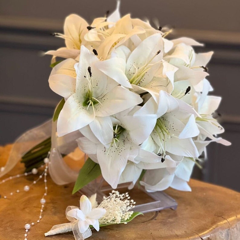 Puede incluir: Un ramo de novia de lirios blancos con tallos y hojas verdes, acentuado con una cinta blanca y una hebra de perlas. Tambi&eacute;n se incluye un prendido a juego con flores blancas y vegetaci&oacute;n.