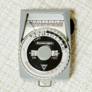 May include: A vintage light meter with a gray body and a circular dial. The dial features numbers and markings in red, white, and black, including the text "ЛЕНИНГРАД 4". A black knob is on the side, and a metal loop is at the bottom.