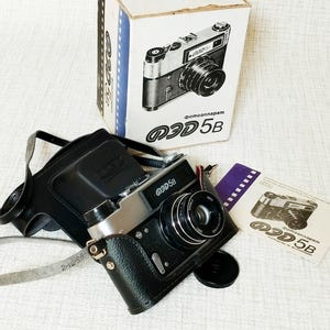 Puede incluir: Una cámara vintage plateada y negra con una funda de cuero, una tapa de lente y un manual de instrucciones. La cámara está junto a su caja original, que presenta un gráfico de la cámara y el texto "Фотоаппарат ЗД5В".