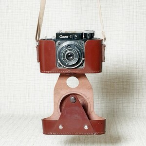 Op de afbeelding: Een vintage zwart-zilveren camera met de tekst "Смена-2" in een bruine leren hoes en riem. De camera is gemonteerd op een bijpassende bruine leren standaard. De cameralens is gecentreerd en de hoes heeft een klassiek ontwerp.