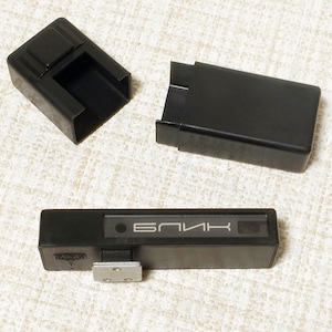 Puede incluir: Kit de accesorios de flash de cámara negro, que incluye una unidad de flash rectangular con la palabra "БЛИК" impresa, una cubierta protectora y un soporte de montaje. El kit está hecho de plástico negro.