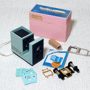 Puede incluir: Un proyector de diapositivas vintage en azul claro y negro, con una caja rosa y varios accesorios. El proyector tiene el texto "ЭКРАН 5". Incluye diapositivas, una bombilla y un manual. La caja mide aproximadamente 20 cm de largo.