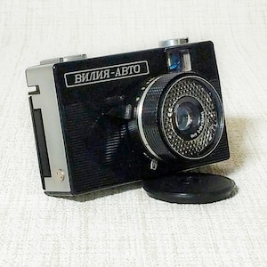 Puede incluir: Una cámara vintage negra y plateada con el texto "ВИЛИЯ-АВТО" en la parte delantera. La cámara tiene una lente prominente y una tapa de lente negra. La cámara está sobre un fondo blanco.