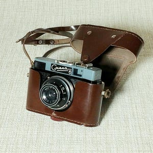 Op de afbeelding: Vintage camera met een bruine leren hoes en riem. De camerabehuizing is grijs en zwart met de Cyrillische tekst "Смена 6" erop. De lens is zwart met zilveren accenten. De hoes is open en toont de camera.