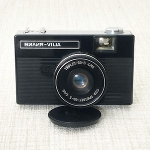 Puede incluir: Una cámara vintage negra y plateada con el texto "ВИЛИЯ-VILIA" en la parte delantera. La lente está etiquetada como "TRIPLET-69-3 4/40". Una tapa de lente negra se encuentra debajo de la lente. La cámara tiene un flash y un botón plateado en la parte superior.