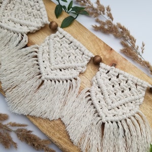 Könnte beinhalten: Drei weiße Makramee-Wandbehänge mit Fransen, jeweils mit einer Holzperle oben.