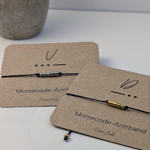 Könnte beinhalten: Zwei Morsecode-Armbänder auf Präsentationskarten. Eines hat silberne Perlen und ein silbernes Rohr, das andere goldene Perlen und ein goldenes Rohr. Beide sind auf schwarzen Kordeln und auf braunen Karten mit dem Text "Morsecode-Armband" und "Fiene Arts" ausgestellt.
