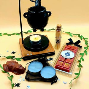 Può includere: Un scaldacera a forma di calderone nero con una base in legno e una candela tea light accesa. Sono inclusi anche un piccolo flacone di vetro di fiammiferi, due candele tea light, una rana di cioccolato e una barretta di cera rossa con glitter dorati. La barretta di cera è etichettata "Unicorn War" con un calderone e una luna crescente.