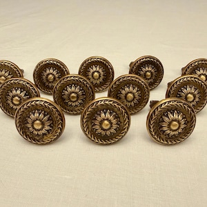 May include: A collection of twelve antique bronze cabinet knobs. Each knob features a central floral design surrounded by a detailed leaf pattern. The knobs are round and have a warm, aged finish, suggesting vintage hardware.