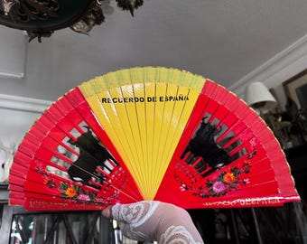 May include: A hand-held fan with a red and yellow design, featuring the text "RECUERDO DE ESPAÑA". The fan has a bull silhouette and floral accents. The fan is a souvenir from Spain.