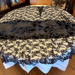 May include: A round tablecloth with a black floral lace overlay. The tablecloth has a black velvet runner and a white underskirt. The lace trim adds a decorative touch to the table setting.