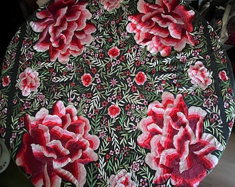 Op de afbeelding: Een zwarte ronde tafelkleed met een geborduurd bloemenmotief. De borduurwerk toont grote roze en rode bloemen met groene bladeren en kleinere roze bloemen overal.