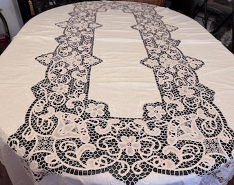 May include: Oval white tablecloth with a black and cream-colored lace design. The lace trim features floral patterns and intricate details, creating an elegant and decorative look. This tablecloth is suitable for special occasions.