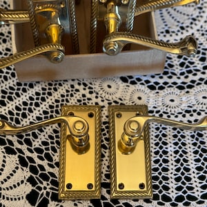 May include: A set of antique brass door handles and plates. The handles feature a curved design with decorative detailing. The plates are rectangular with a rope-like border. The hardware is displayed on a white lace tablecloth.