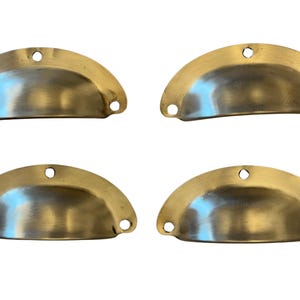 Peut inclure: Quatre poignées en forme de coupe de couleur laiton antique, disposées en carré. Chaque poignée a une forme semi-circulaire avec trois trous pour le montage. Les poignées ont un aspect légèrement vieilli.