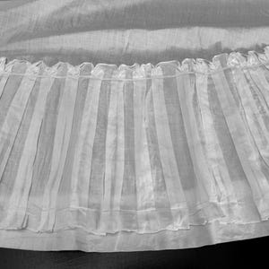 May include: A white sheer fabric with a ruffled edge and vertical pleats. The fabric is translucent and has a delicate, airy feel.