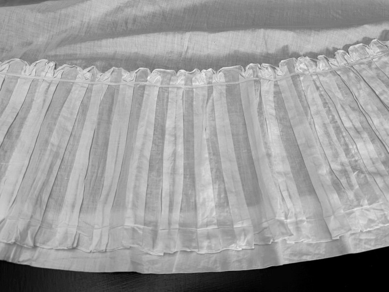 May include: A white sheer fabric with a ruffled edge and vertical pleats. The fabric is translucent and has a delicate, airy feel.