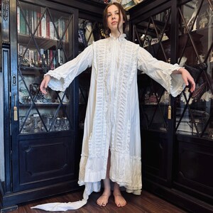 May include: A white, long-sleeved, lace-trimmed gown with a high collar and a ruffled hem. The gown is made of a lightweight fabric and has a flowing silhouette.