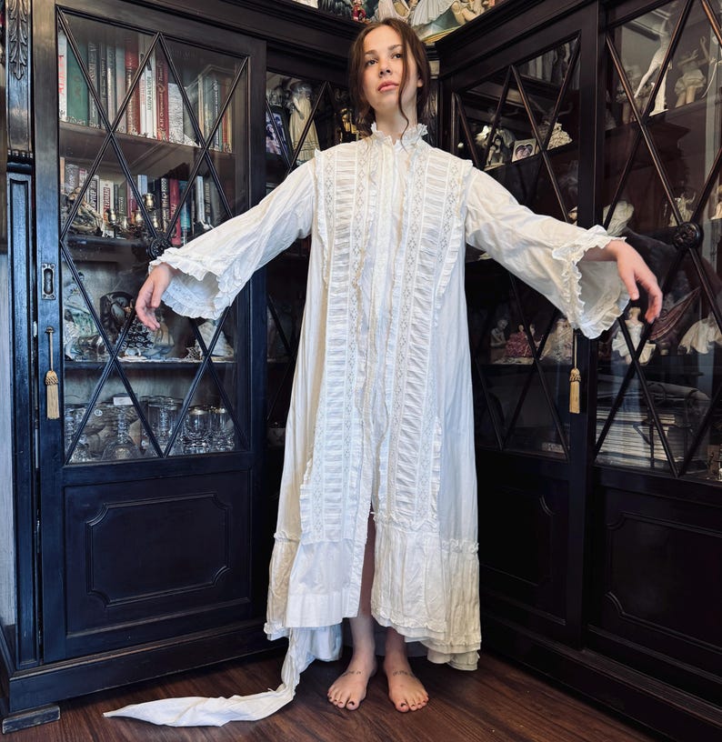 May include: A white, long-sleeved, lace-trimmed gown with a high collar and a ruffled hem. The gown is made of a lightweight fabric and has a flowing silhouette.