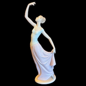 Op de afbeelding: Een porseleinen beeldje van een ballerina in een sierlijke pose. Het beeldje is geschilderd in zachte pastelkleuren, met een lichtblauwe lijfje en een vloeiende roze rok. De arm van de ballerina is geheven en ze kijkt omhoog.