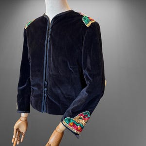 Puede incluir: Chaqueta de terciopelo azul marino oscuro con detalles bordados coloridos en los hombros y los puños. La chaqueta tiene cuello redondo, mangas largas y pequeños botones en la parte delantera. El bordado presenta detalles rojos, verdes y dorados.