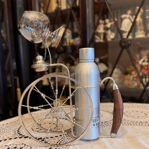 May include: Silver bottle holder with a decorative wheel design, a glass decanter, and a silver bottle. The holder has a wooden handle.