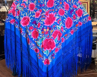 Peut inclure: Un châle bleu vif orné d'un motif floral dans des tons de rose, rouge et vert. Le châle présente une bordure bleu foncé avec des détails au crochet et de longues franges. Le design est riche et détaillé.