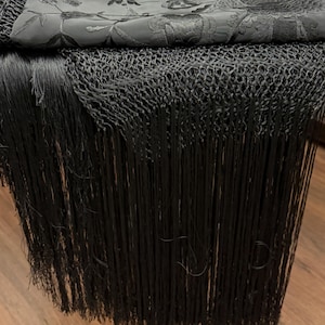 May include: A black shawl with detailed floral embroidery and long fringe. The top layer has a detailed floral pattern, while the bottom layer has a woven net design with long, flowing fringe. The shawl is draped over a wooden surface.