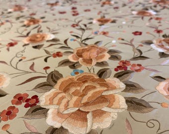 Op de afbeelding: Een lichtbeige stof met een ingewikkeld geborduurd bloemenpatroon. Het ontwerp bevat grote, gelaagde perzik- en beige bloesems, kleinere rode bloemen en donkerbruine bladeren, verbonden door dunne, kronkelende donkerbruine stengels.