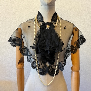 May include: A vintage-style black lace collar and capelet with floral embroidery. The capelet features a ruffled front and is adorned with a pearl necklace and a decorative brooch. The garment is displayed on a mannequin.