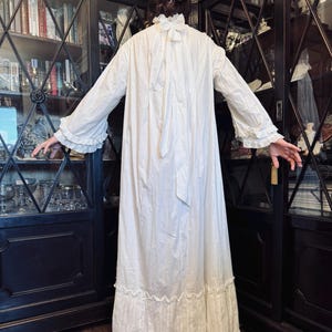 May include: A white cotton nightgown with a ruffled neckline and long sleeves. The gown has a gathered skirt and a tie at the back of the neck.