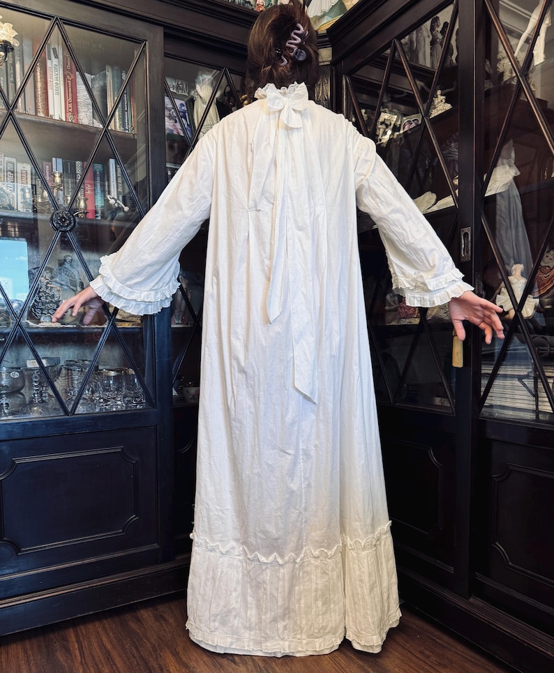 May include: A white cotton nightgown with a ruffled neckline and long sleeves. The gown has a gathered skirt and a tie at the back of the neck.