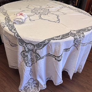 May include: A white, round tablecloth with detailed lace edging and a floral pattern in the centre. A rolled white napkin with a pink ribbon sits on the cloth. The tablecloth is draped over a wooden table.