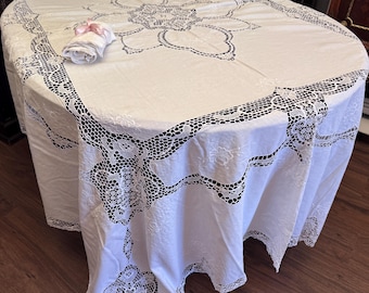 May include: A white, round tablecloth with intricate lace detailing around the edges and a floral design in the center. A rolled-up white napkin with a pink ribbon rests on the tablecloth. The tablecloth is draped over a wooden table.