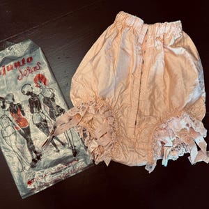 May include: Vintage peach-colored bloomers with lace trim and a matching package. The package has the text "conjunto Soras" and illustrations of women in lingerie. The bloomers have a button closure.