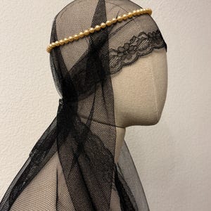 May include: A black tulle veil with a delicate lace trim and a gold beaded headband. The veil is draped over a mannequin head, showcasing the intricate details of the fabric and design.