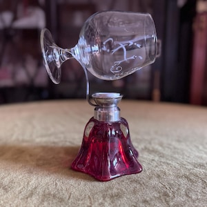 Può includere: Un bicchiere da vino in vetro trasparente con un design decorativo è tenuto da un supporto in metallo argentato sopra una base in vetro rosso rubino. La base ha una forma svasata a campana. L'oggetto è su una superficie dorata.
