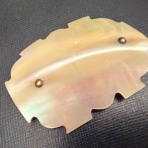 May include: A pearlescent, leaf-shaped decorative object with two small, gold-colored rivets. The object has a scalloped edge and a subtle iridescence, reflecting a range of colors. The item is likely a decorative element or embellishment.