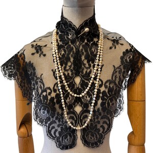 May include: A black lace collar featuring a floral pattern with scalloped edges, worn over a mannequin. A long, double-strand necklace made of off-white, round pearls hangs over the lace. The collar is secured at the neck with a small, round pin.