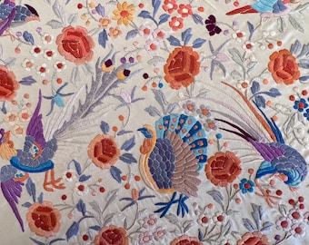 Peut inclure: Un tissu blanc avec des oiseaux et des fleurs brodés de couleurs vives. Les oiseaux sont stylisés et ont de longues queues flottantes. Les fleurs sont dans des tons de rose, rouge et jaune.