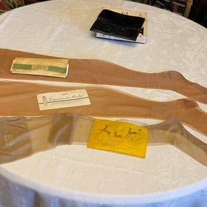May include: Three pairs of sheer, tan-coloured stockings laid out flat. Each pair includes a branded paper label, with one labelled Balkis, one Eugenia de Montijo, and one featuring a yellow graphic print with the text Unline.