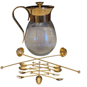 May include: A clear glass jug with gold-coloured handle, rim, and lid, and etched floral designs. Several gold-coloured bar spoons and forks are arranged around the jug, suggesting a barware set.