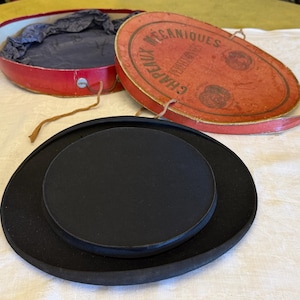 May include: A black top hat with a flat crown and wide brim, accompanied by an oval red storage box. The box lid features the text CHAPEAUX MECANIQUES PERFECTIONNES and includes a thin string closure and a dark blue fabric interior lining.