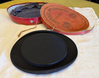 May include: A black top hat with a flat crown and wide brim, accompanied by an oval red storage box. The box lid features the text CHAPEAUX MECANIQUES PERFECTIONNES and includes a thin string closure and a dark blue fabric interior lining.
